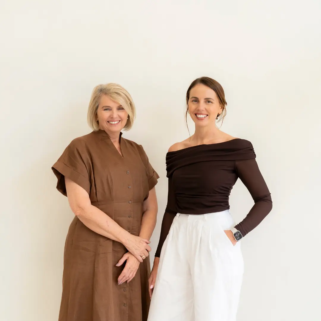 Two smiling women standing in front of a plain light background, one in a brown dress and the other in a dark off-shoulder top with white pants.
