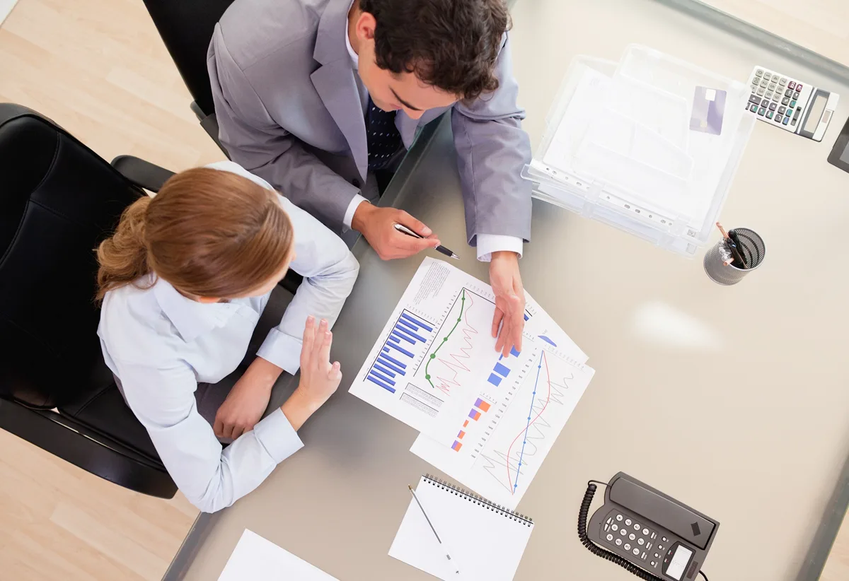High-angle view of a man and woman in business attire discussing printed financial charts at a desk.