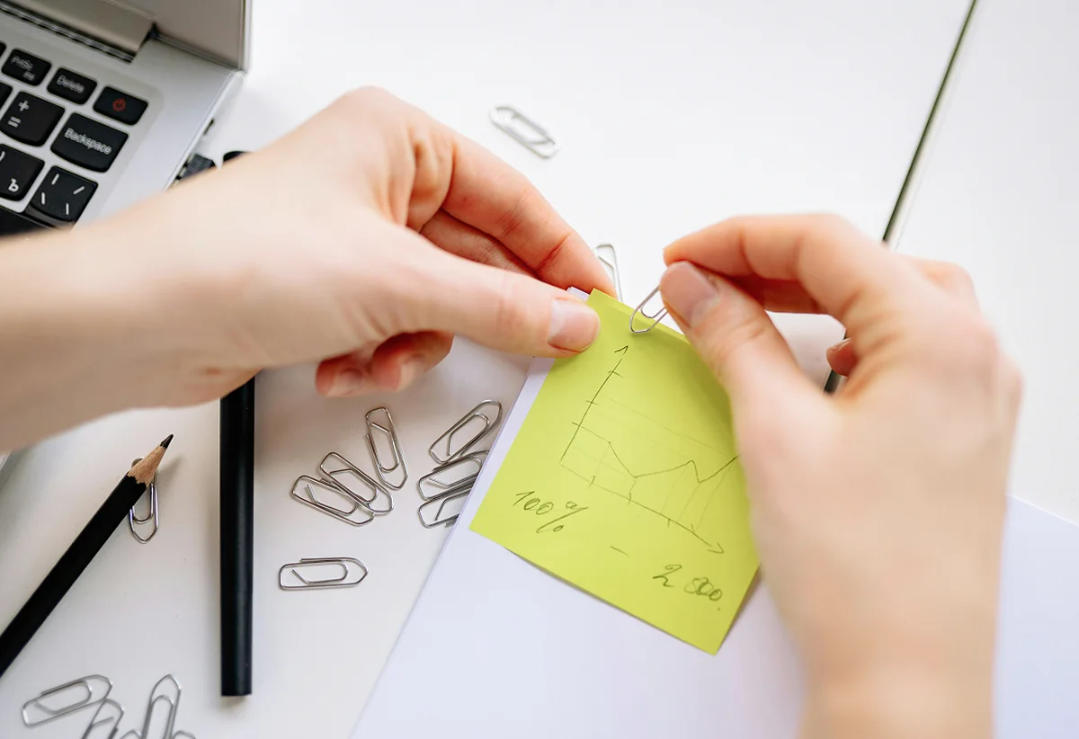 Hands using a paperclip to attach a yellow sticky note with a hand-drawn line graph onto a notebook page.