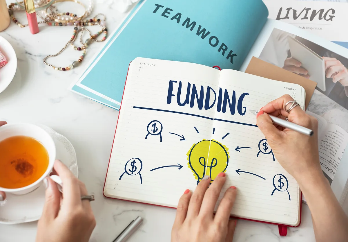 Overhead view of hands writing the word "Funding" in a notebook surrounded by a teacup and business documents.