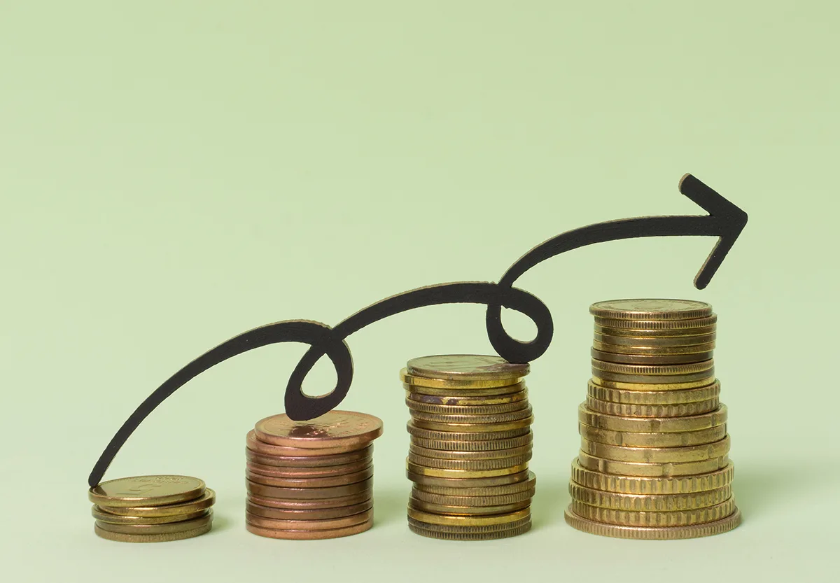 Four ascending stacks of gold and copper coins under a black wavy arrow pointing upwards to represent financial progress.