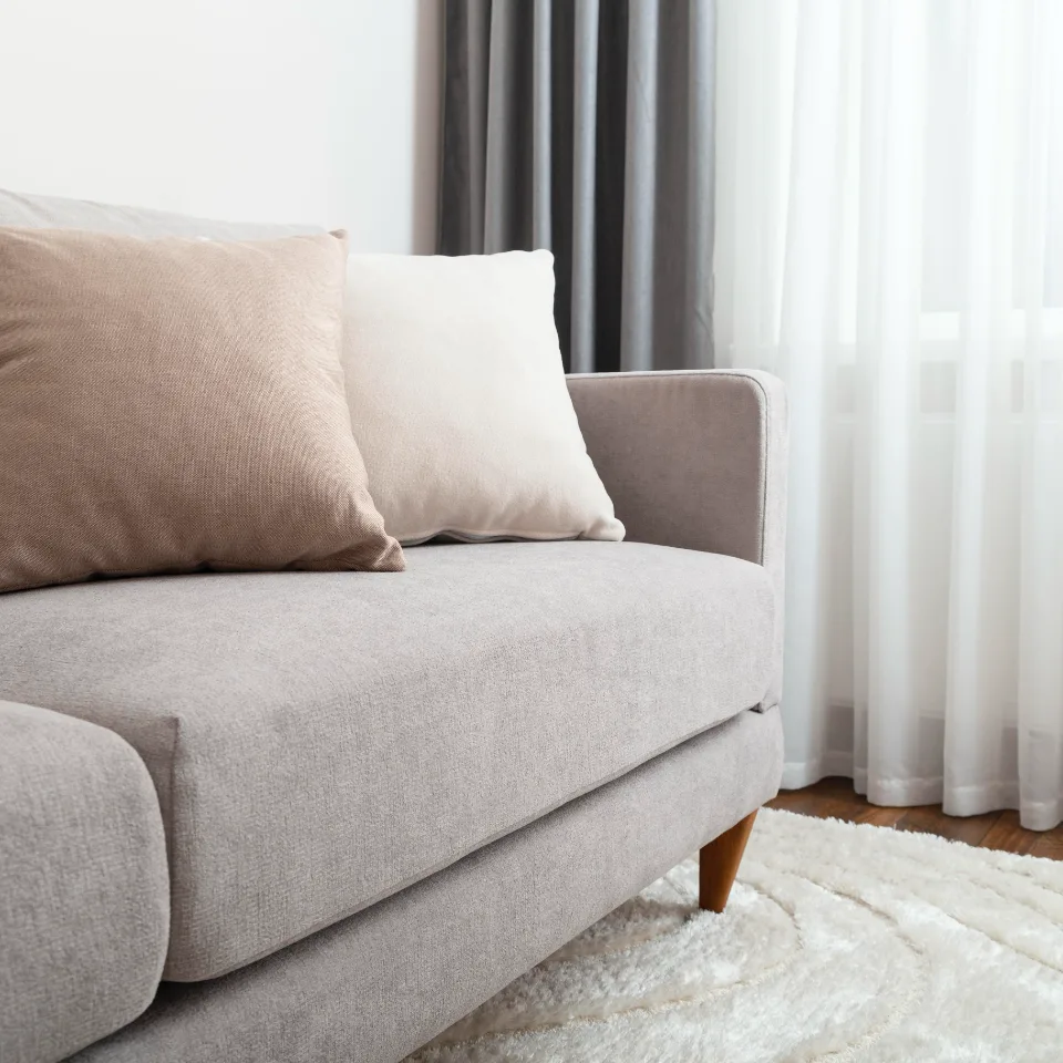 Light grey fabric sofa with beige cushions beside a window with curtains.