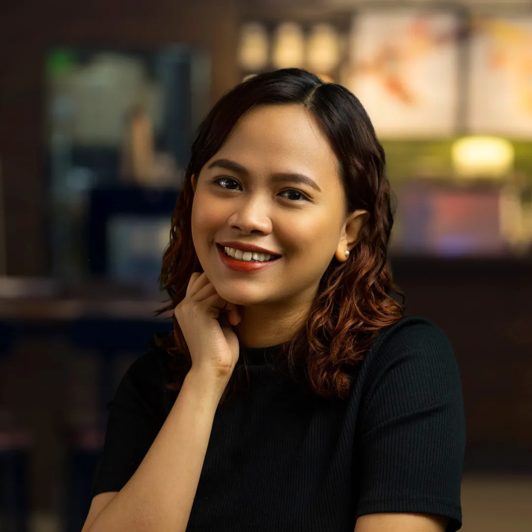 Smiling woman with wavy brown hair wearing a black top and resting her hand on her neck in a softly lit indoor setting.