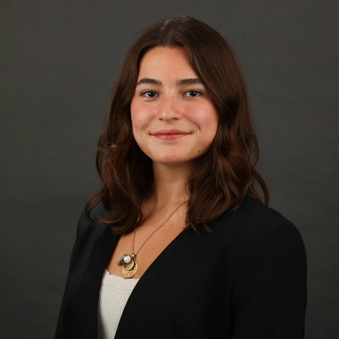 Smiling woman with shoulder-length brown hair wearing a black blazer and gold necklace against a dark background.
