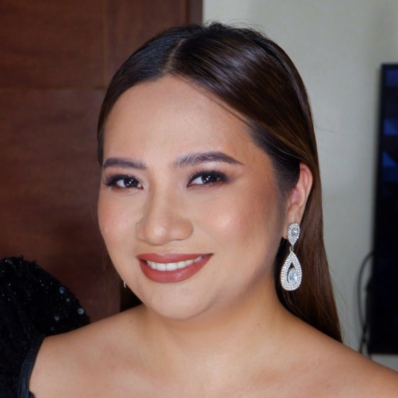Woman with long brown hair and elegant makeup smiling, wearing large teardrop-shaped earrings and a black off-shoulder top.