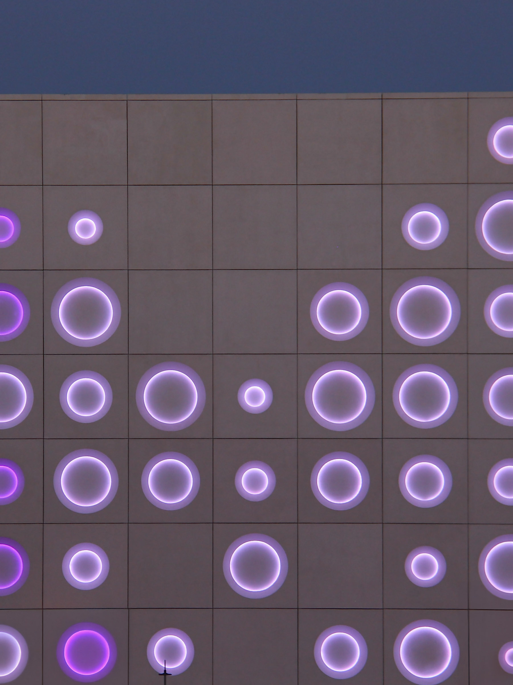 Night elevation with concentric circular lighting pattern across GRC panels viewed from street level.