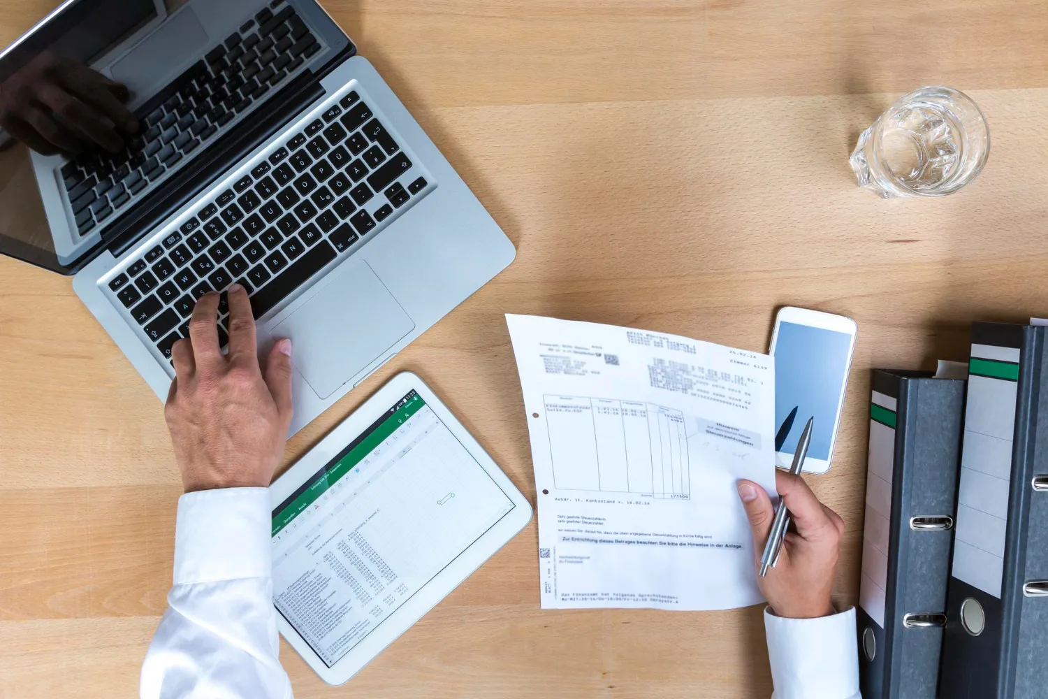 Person working at wooden desk with laptop, tablet, documents, and office supplies overhead view