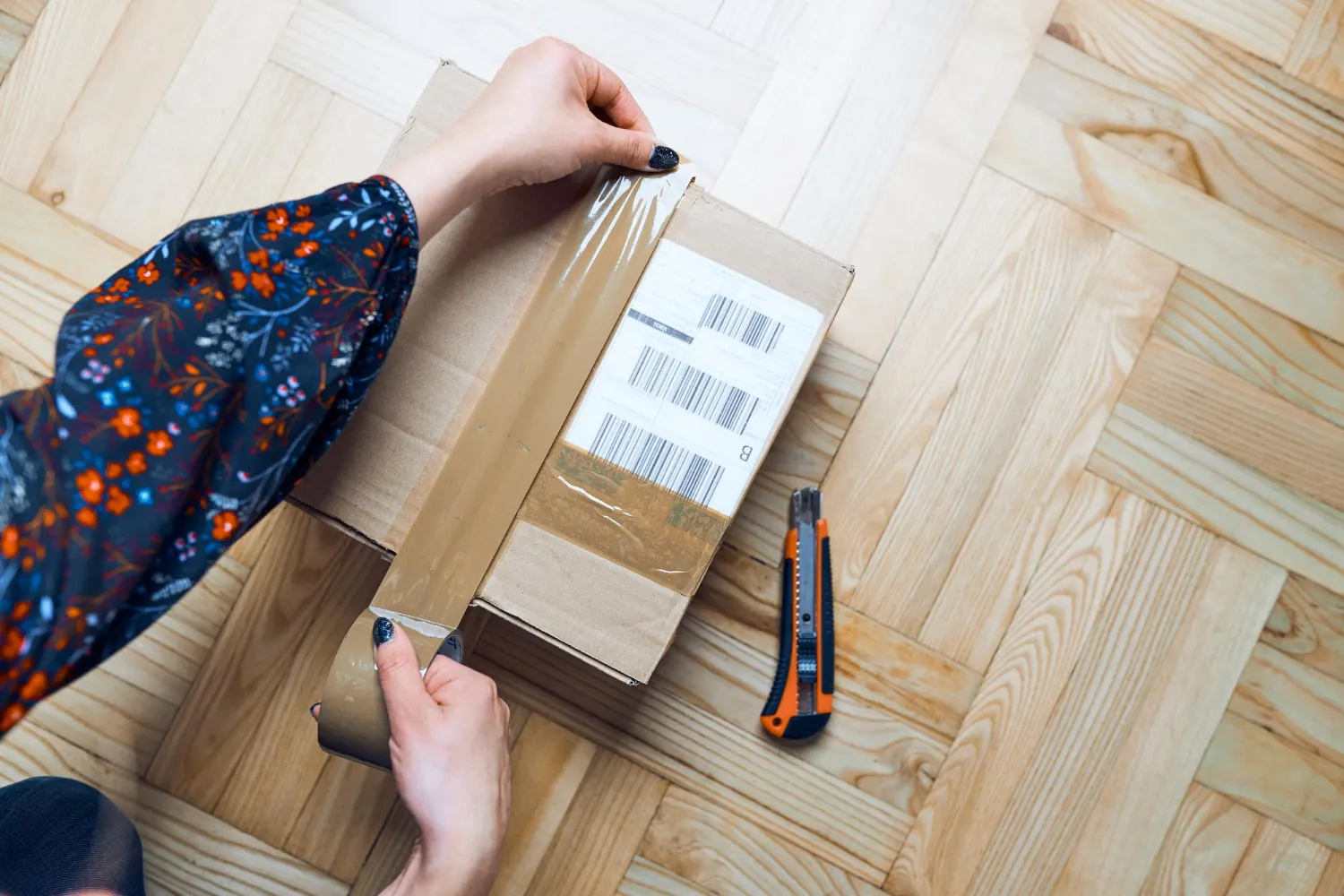 Person opening cardboard delivery box with barcode labels on wooden floor.