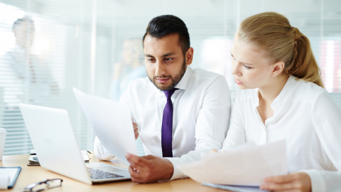 Two colleagues comparing printed invoices against figures on a laptop screen.