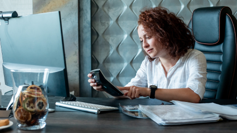 Business owner checking a tablet at a desk stacked with financial paperwork.