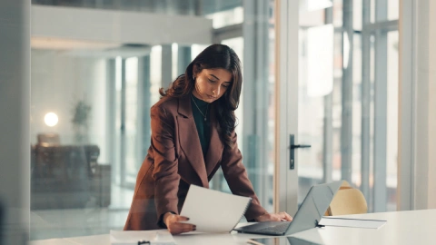 Businesswoman comparing printed account documents against a laptop at a standing desk.