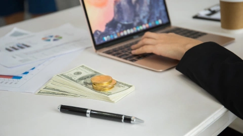 Cash and gold coins beside a laptop and financial charts on a desk.