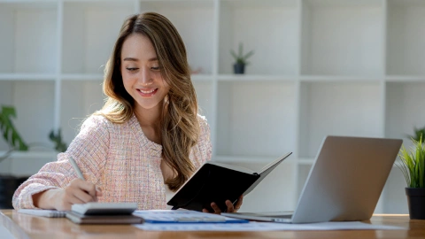Businesswoman calculates costs with a notebook and calculator at her desk.