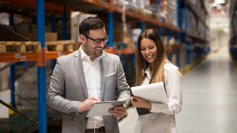 Two colleagues review inventory on a tablet in a warehouse aisle.