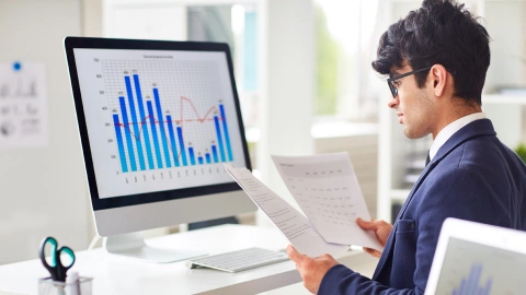 Analyst compares printed reports against bar charts on a desktop monitor.