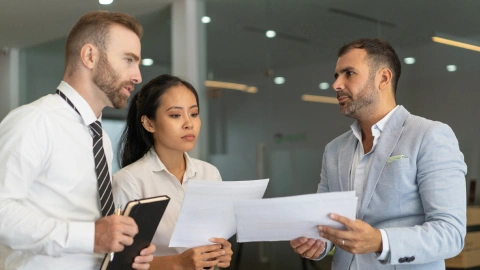 Three professionals closely review contract documents together in an office.