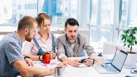 Three coworkers reviewing a phone together at a table with a laptop.