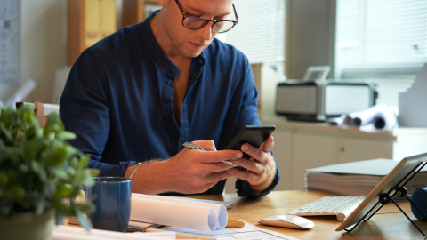 Man checking his phone at a desk with rolled documents nearby.