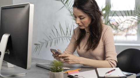 Woman checking her phone at a desk with a desktop computer.
