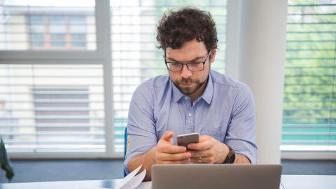 Man checking his phone at a desk with a laptop open.