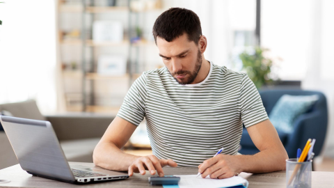 Man using a calculator and taking notes at a laptop in a home office.