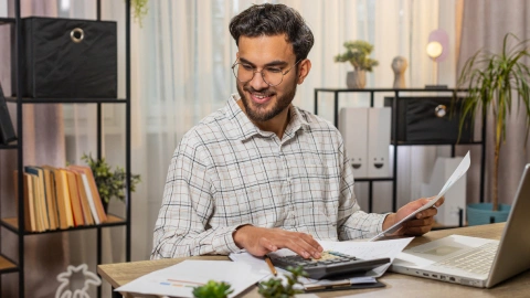 Man smiling while using a calculator with a laptop at his desk.