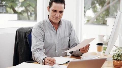 Man reviewing a document at his desk beside a laptop.