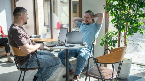Two freelancers talk over laptops at a sunlit café table