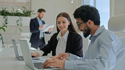 Two coworkers research options on laptops in a bright office