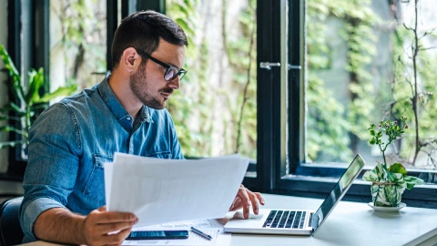 Man reviewing documents beside an open laptop near a window.