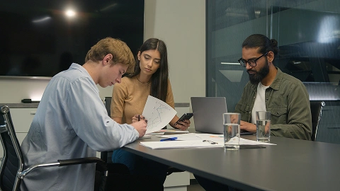 Three coworkers working together at a table with a laptop and papers.