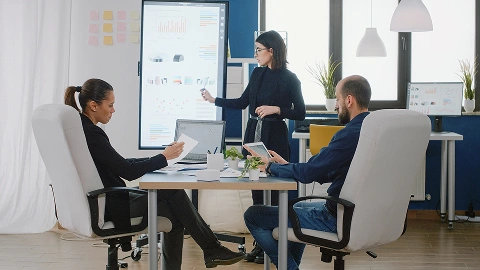 Woman presenting data charts on a screen to two seated colleagues.