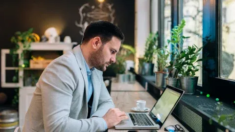 Man in a suit working on a laptop near a window with plants.