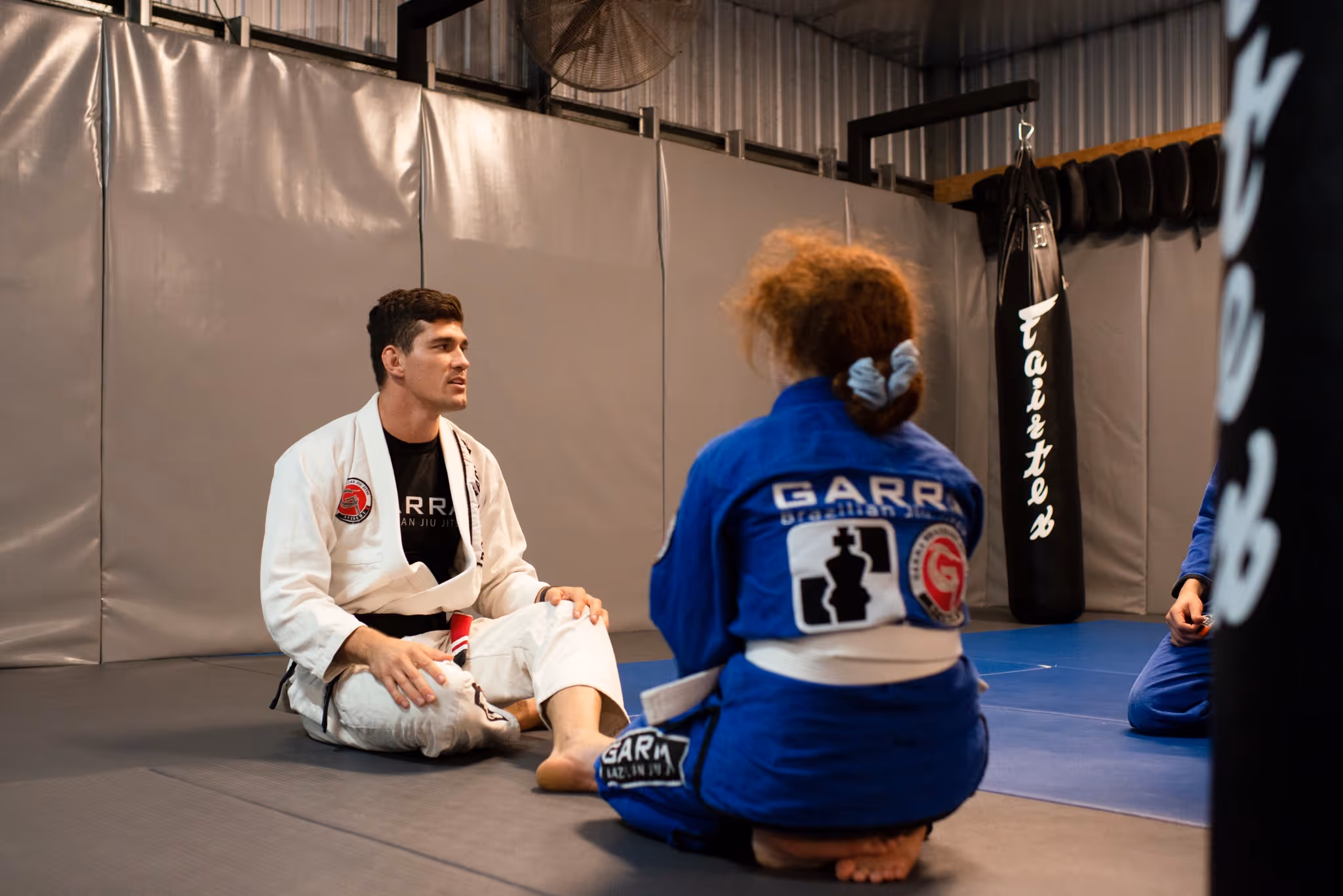 Two people in Brazilian Jiu-Jitsu gis sitting on mats in a martial arts gym, with a punching bag in the background.