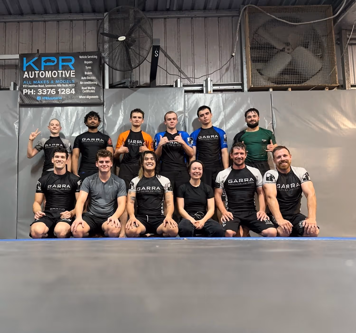 Group of eleven people in martial arts attire posing on a mat inside a gym with a KPR Automotive sign and two large fans on the wall behind them.