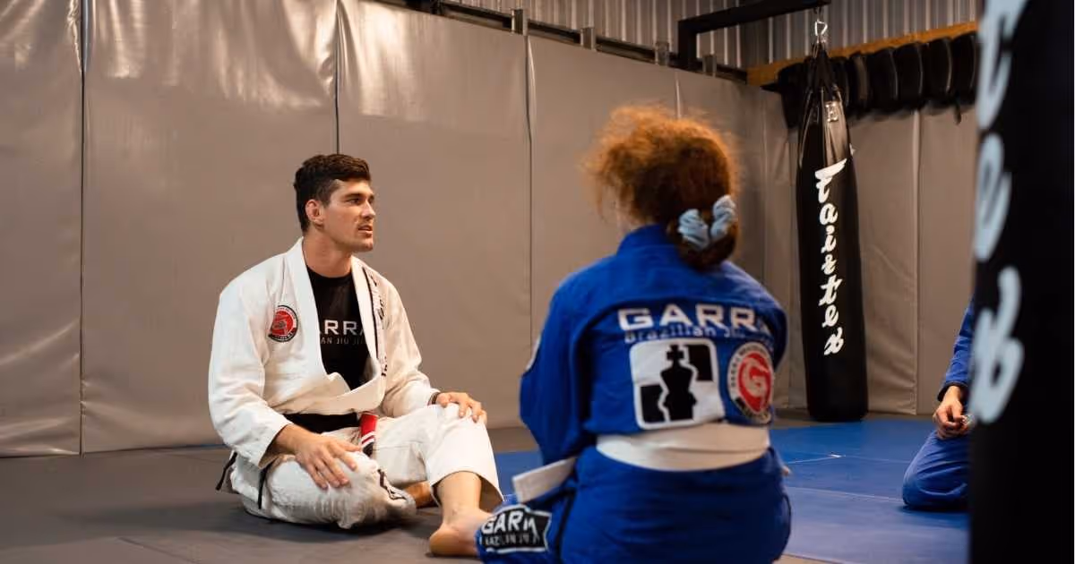 Two martial artists in gis seated on a mat inside a training gym, with one instructor speaking to a student.