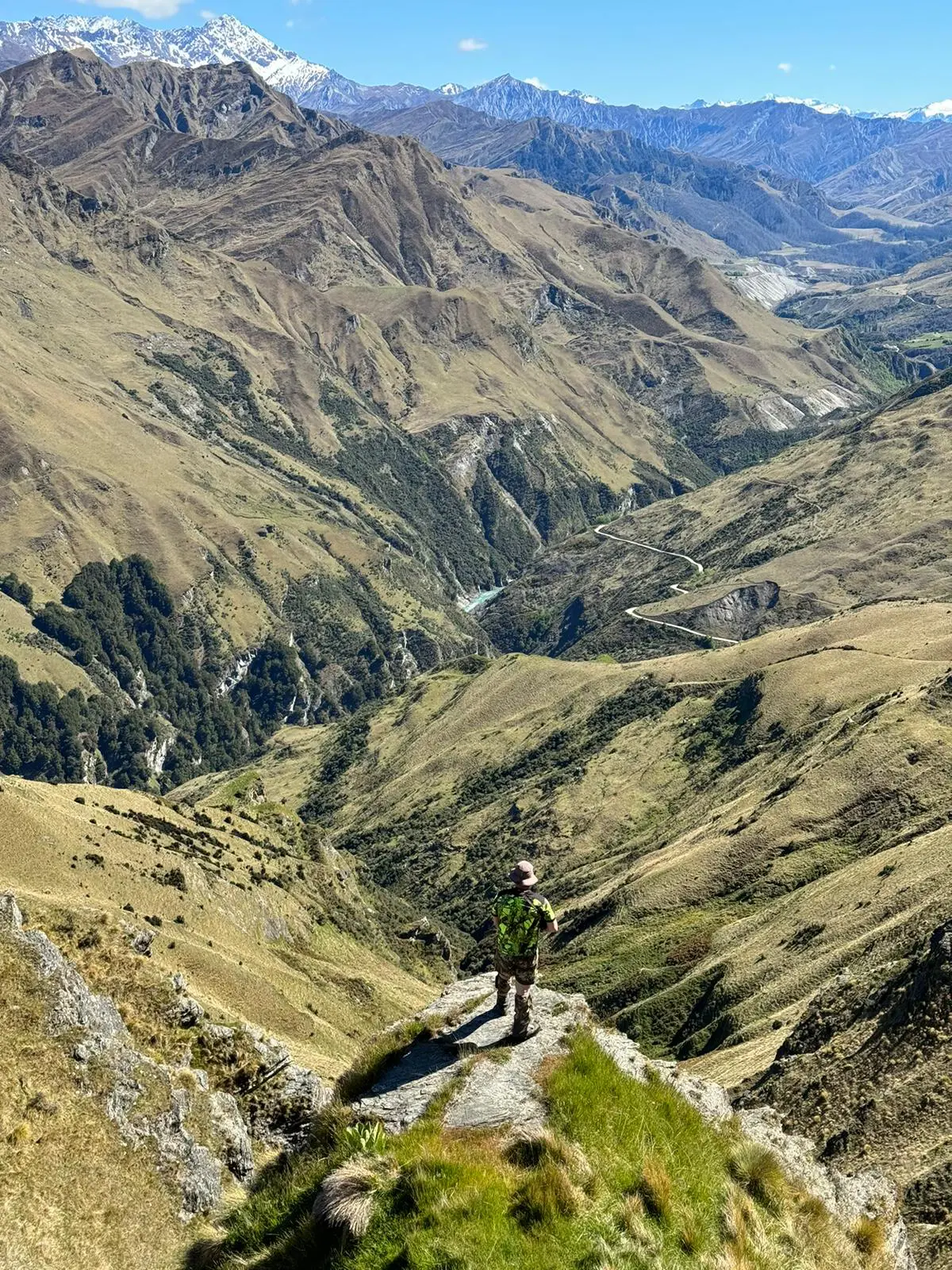 Person standing on a grassy hilltop overlooking a winding road through a mountainous landscape with snow-capped peaks in the distance.