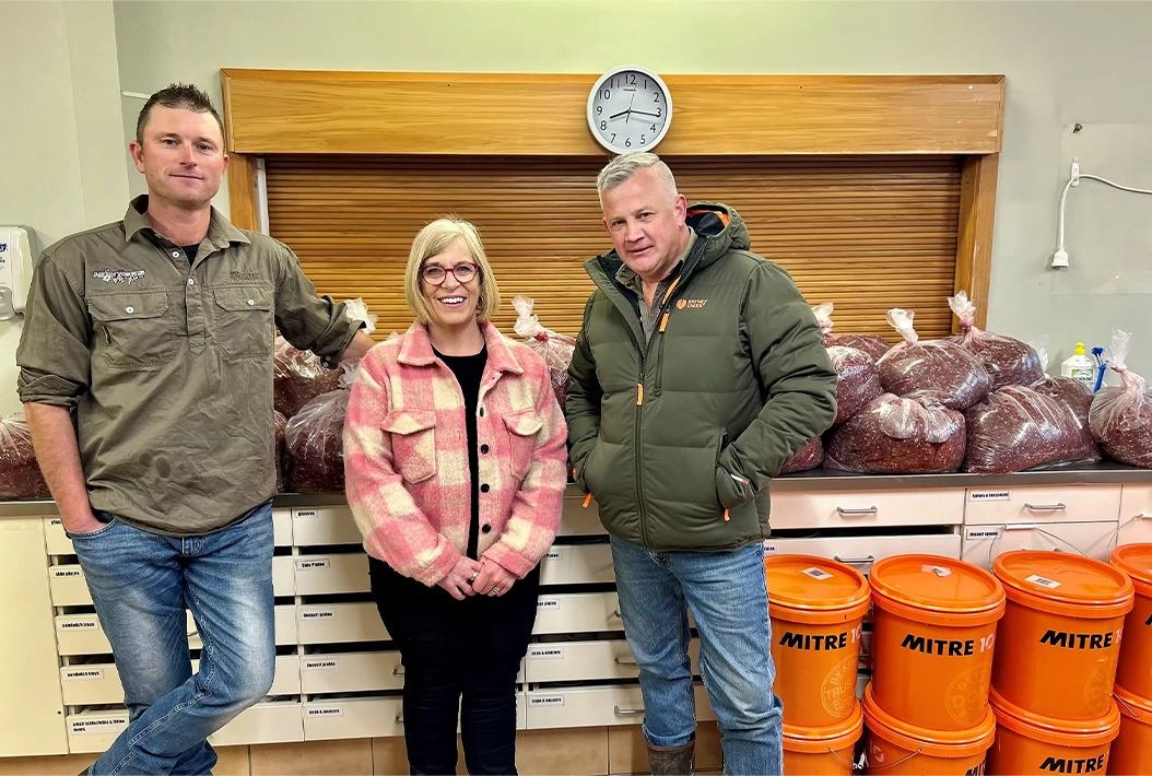 Three people standing indoors in front of a counter with large bags of red beans and stacked orange Mitre 10 buckets.