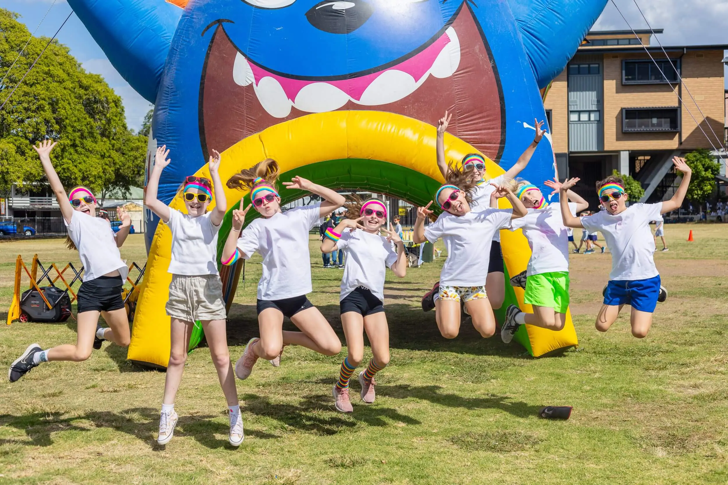 Group of eight children wearing white shirts, colorful headbands, and sunglasses jumping energetically in front of a large inflatable blue character with a big smile.