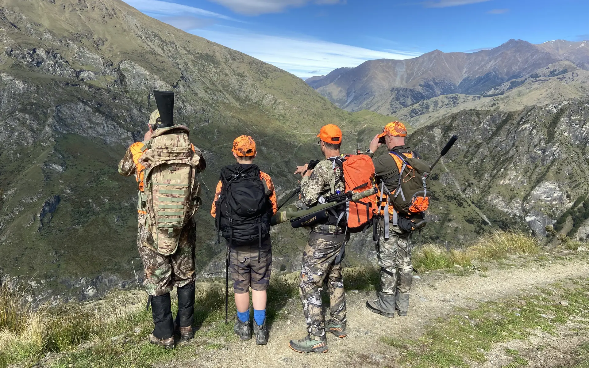 Four hunters in camouflage and orange gear stand on a mountain trail overlooking a valley with rugged hills under a clear blue sky.