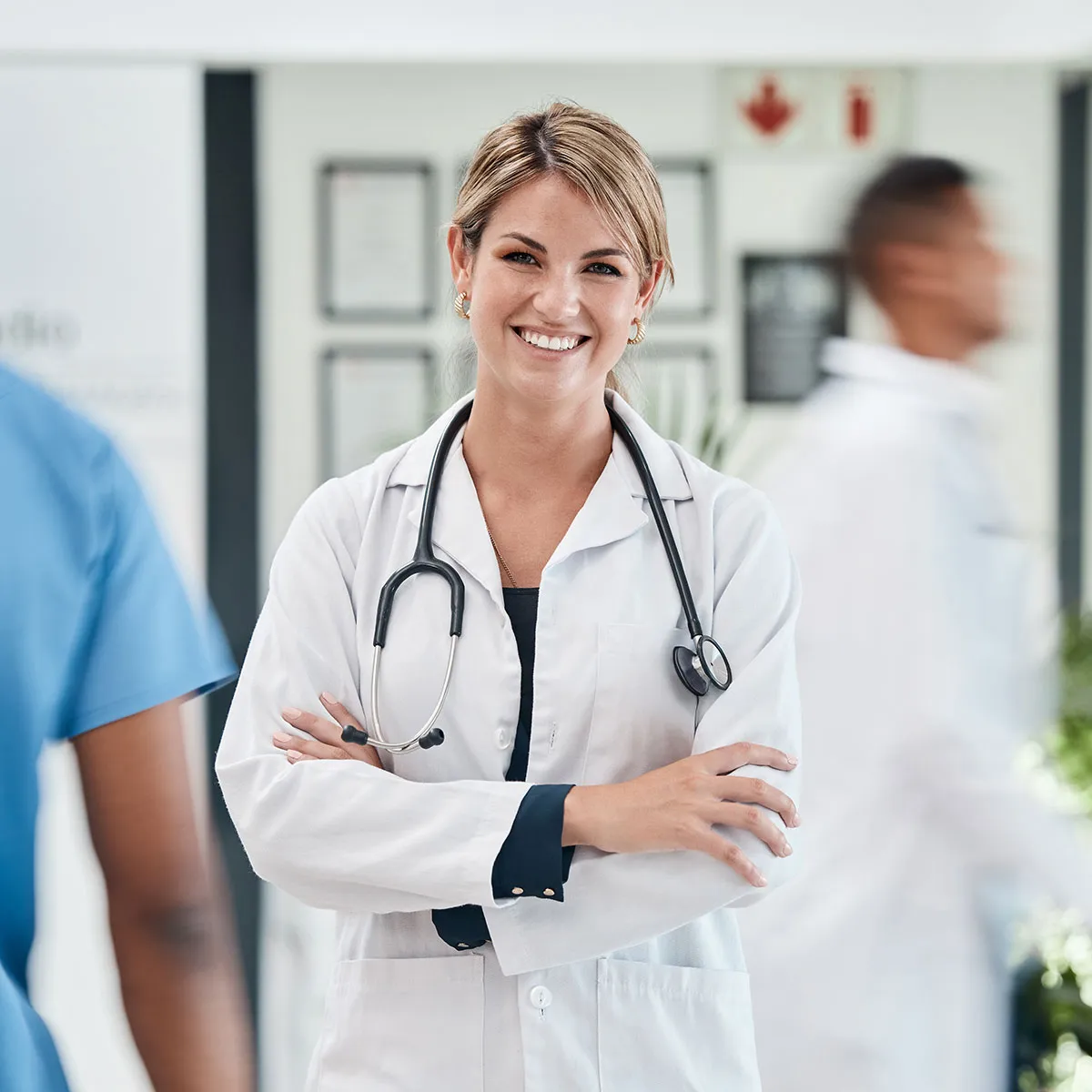 Doctor in a white coat with a stethoscope, standing with arms crossed in a medical facility. Other medical staff are visible in the background.