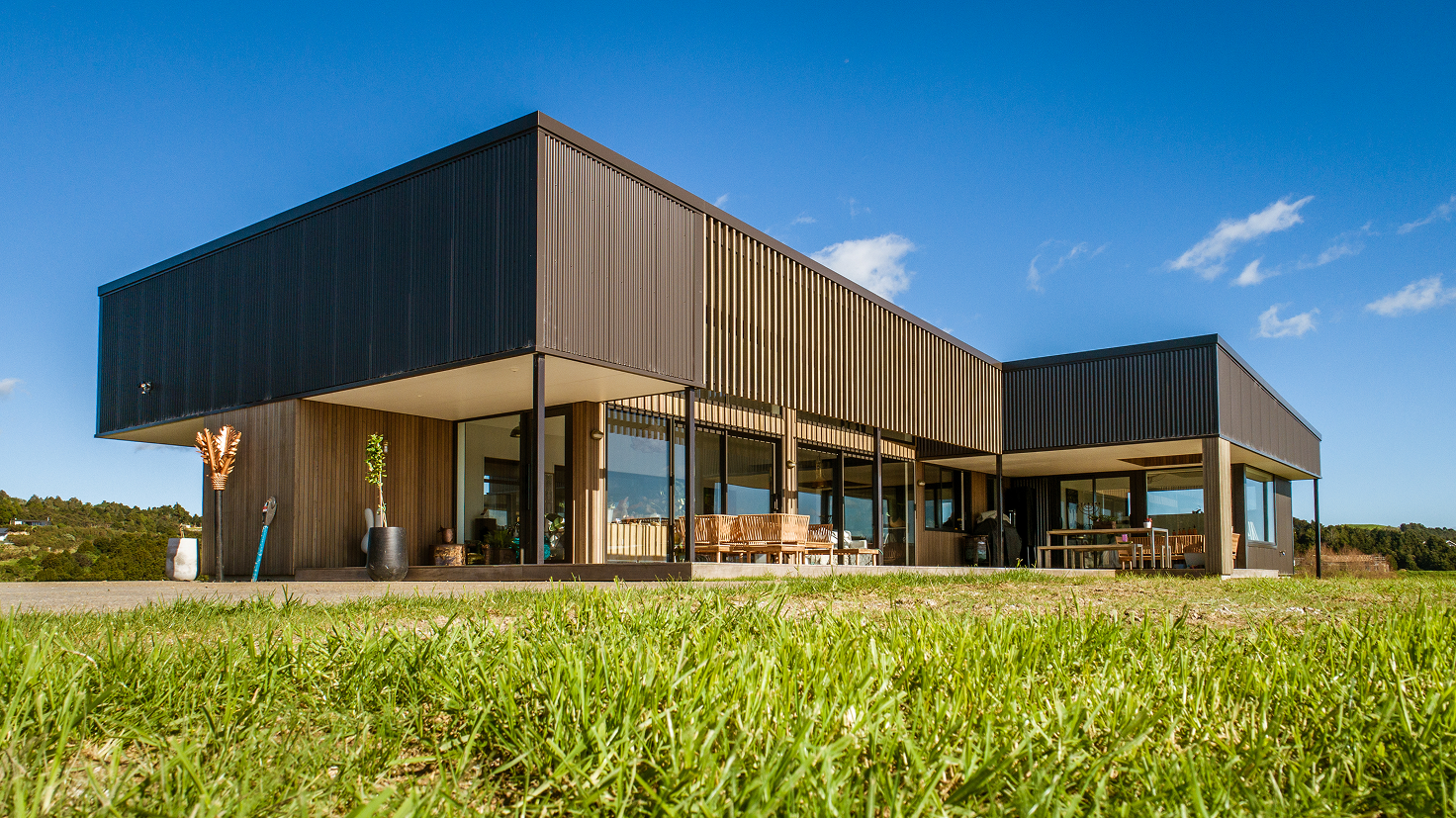 Modern single-story house with large glass windows and wooden panels surrounded by green grass under a clear blue sky.