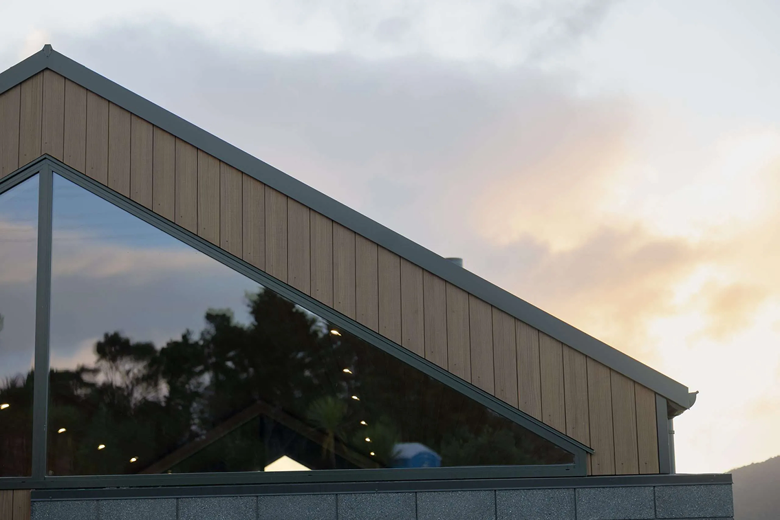 Modern building facade with large triangular glass window reflecting trees and sky at sunset.