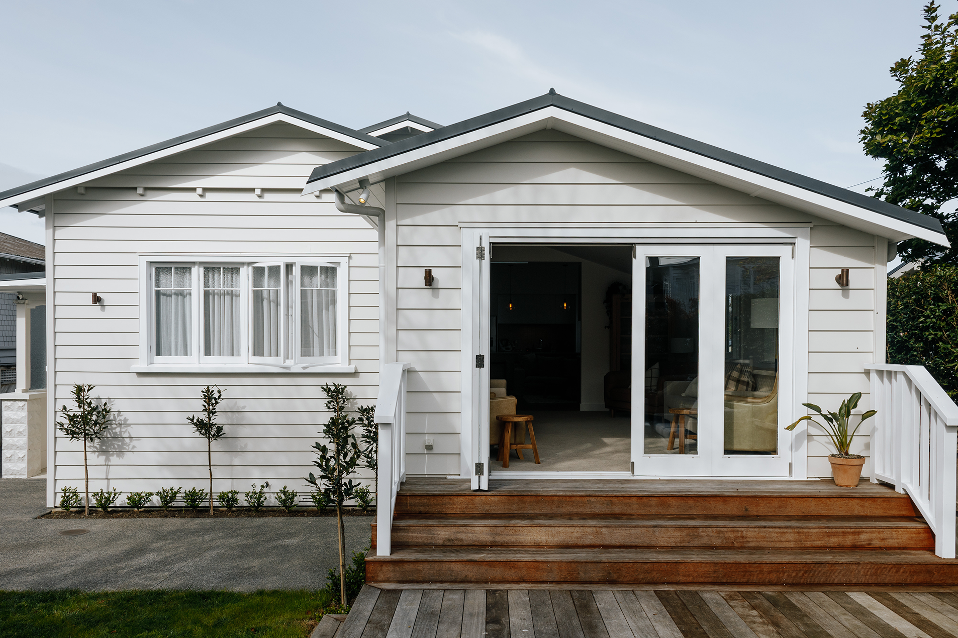 White modern house exterior with open glass doors, wooden steps, small trees, and a potted plant on the porch.