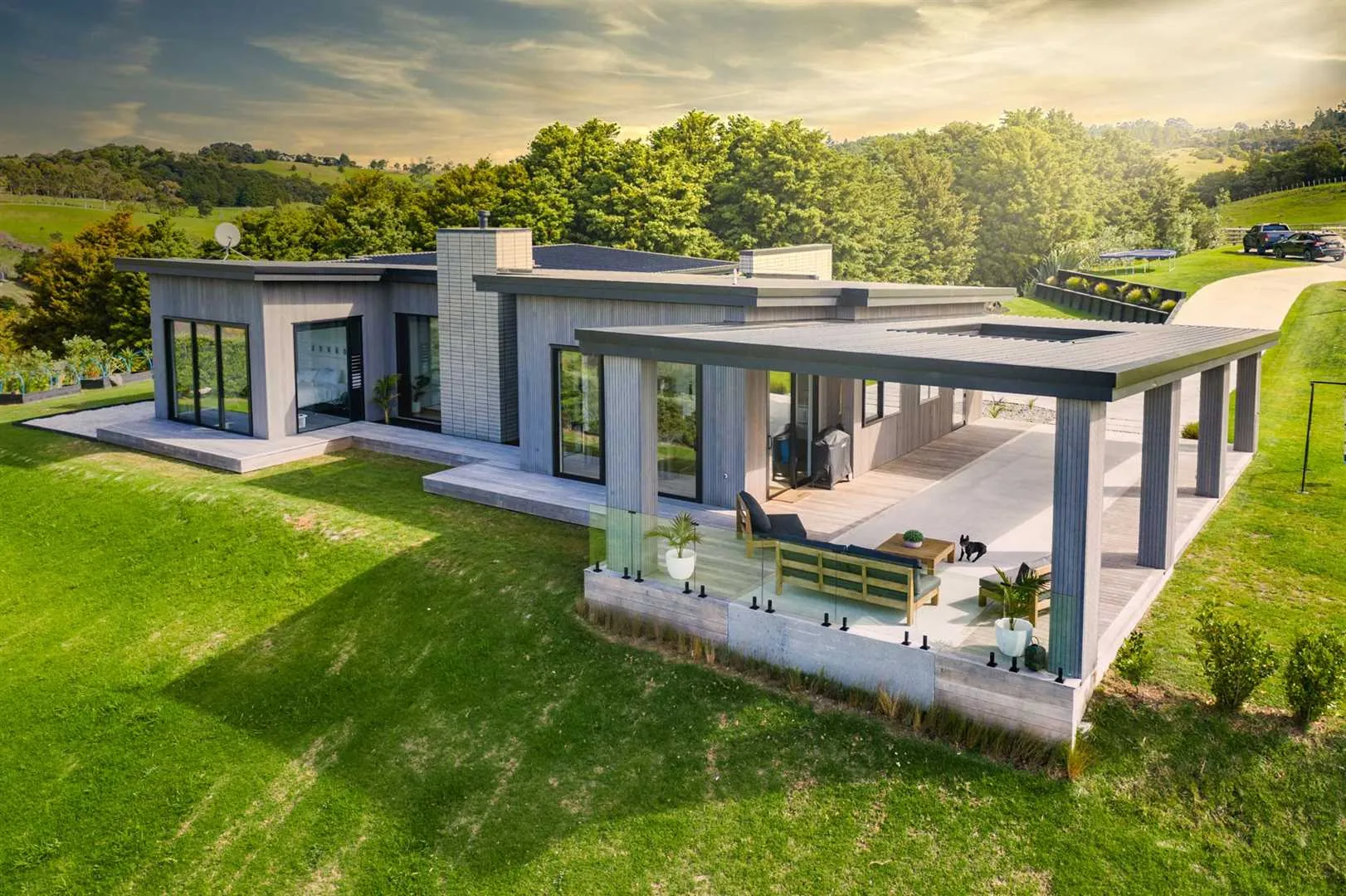 Modern single-story house with large glass windows, a covered patio with outdoor furniture, and a green lawn surrounded by trees under a partly cloudy sky.