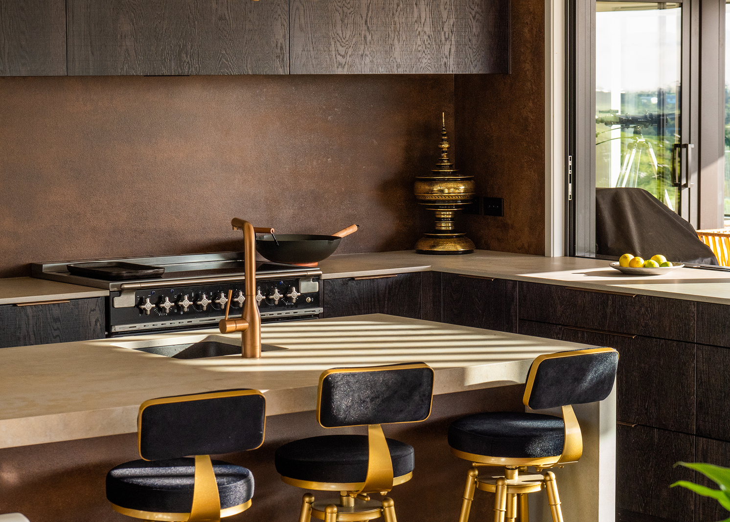 Modern kitchen with beige countertop island featuring a bronze faucet, black and gold bar stools, and a cooking range with a wok pan, illuminated by sunlight.