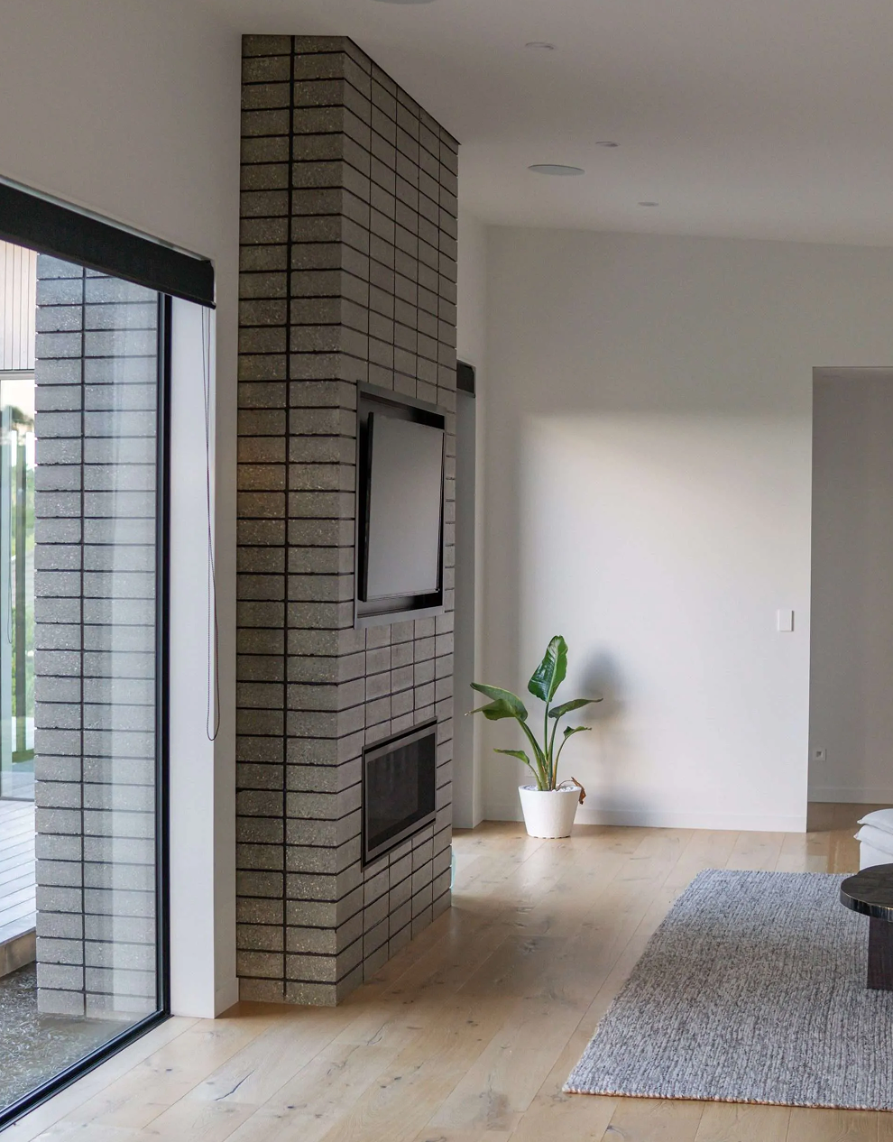 Modern living room with a brick fireplace wall featuring a mounted TV, a potted plant near the corner, and a large window with a black roller blind.