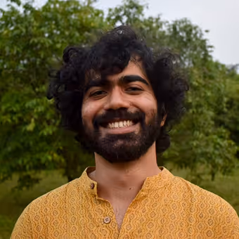 Smiling man with curly hair and beard wearing a yellow patterned shirt outdoors with green foliage background.