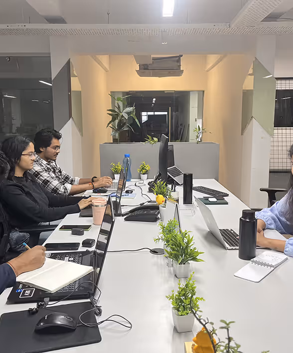 People working at a long office table with laptops, notebooks, and small potted plants in a modern workspace.