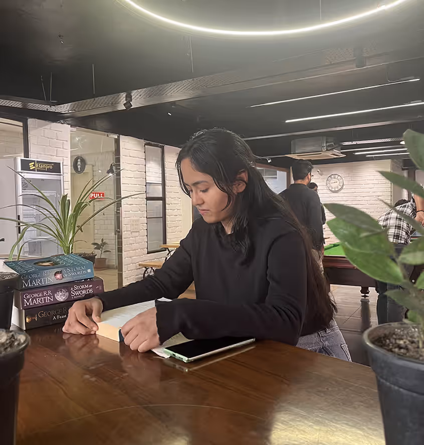 Young woman with long dark hair reading a book at a wooden table in a modern room with plants and a stack of George R.R. Martin books nearby.